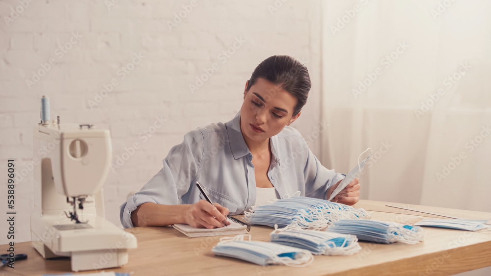 Seamstress holding medical mask and writing on notebook near sewing machine. Seamstress holding medical mask and writing on notebook near sewing machine.