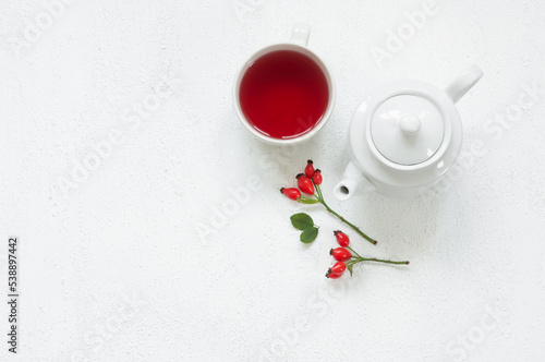 Rosehip glass cup of herbal tea medicinal plants with rosehip branch and ceramic teapot on white table. Dogrose winter tea background