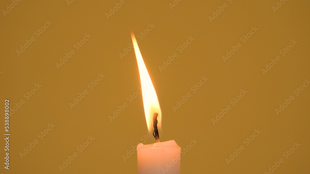 Close-up of a single wax candle burning. Isolated glowing candle against a yellow background. Background or illustration of religious ceremony, spiritual memory, faith and traditions. Camera zoom out. Close-up of a single wax candle burning. Isolated glowing candle against a yellow background. Background or illustration of religious ceremony, spiritual memory, faith and traditions. Camera zoom out.