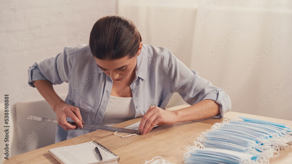 Brunette designer measuring medical mask near notebook in atelier. Brunette designer measuring medical mask near notebook in atelier.