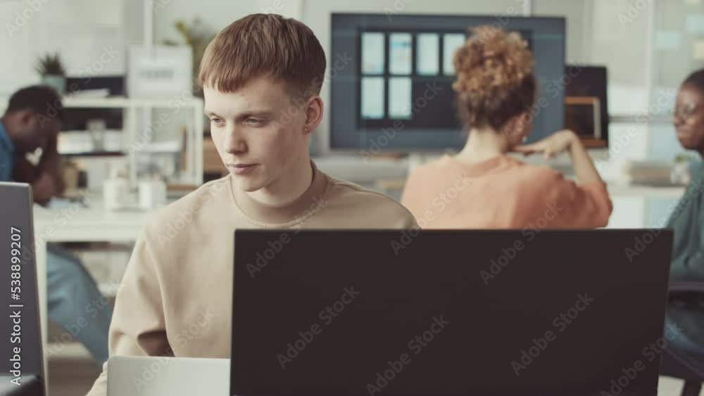 Young Caucasian male programmer looking at computer monitor while working in IT office with diverse colleagues Young Caucasian male programmer looking at computer monitor while working in IT office with diverse colleagues