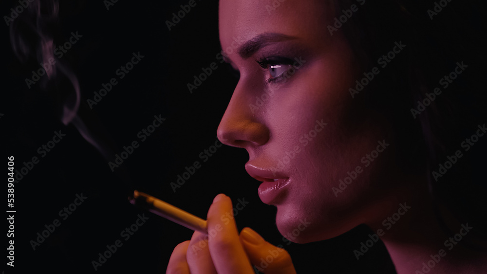 Obraz premium Side view of young woman holding blurred cigarette on black background.