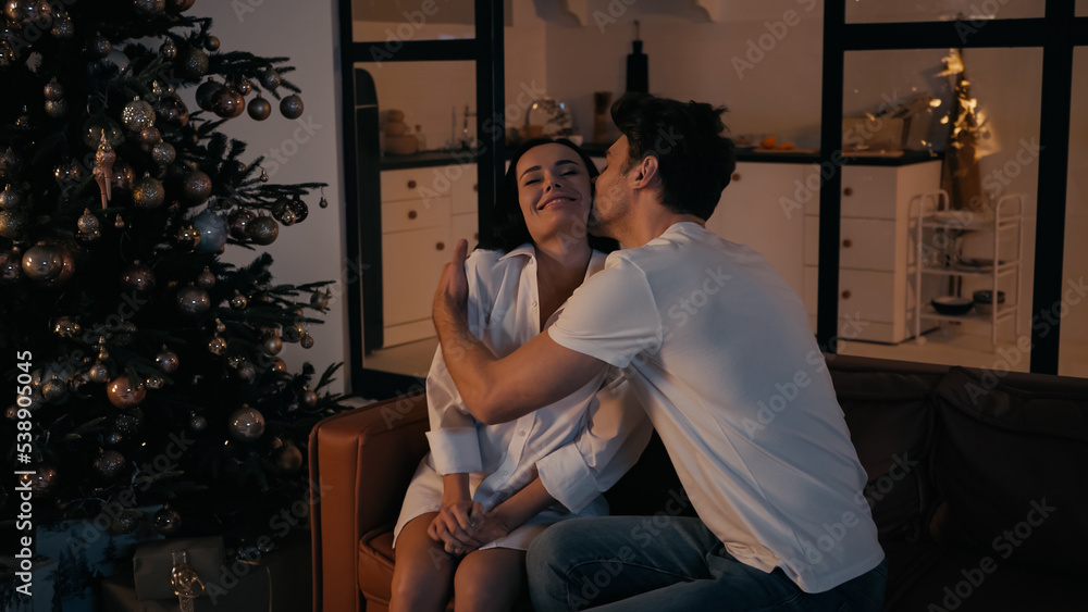 man kissing cheek of happy woman sitting on couch near christmas tree.