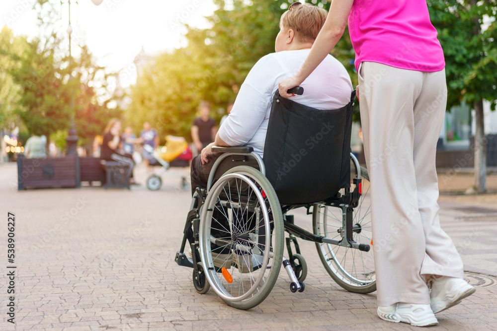 Back view of young woman helping mature woman in wheelchair in the city Back view of young woman helping mature woman in wheelchair in the city