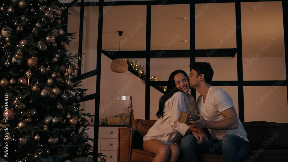 man holding present and kissing cheek of happy woman near christmas tree.