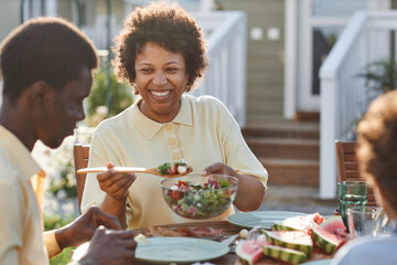 Portrait of smiling African American woman serving food to family while enjoying dinner together outdoors in sunlight