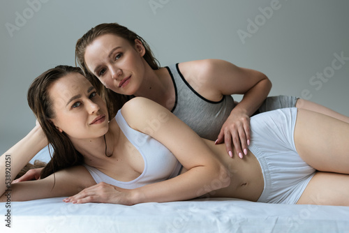 sexy couple of lesbians lying on bed in underwear and looking at camera isolated on grey.