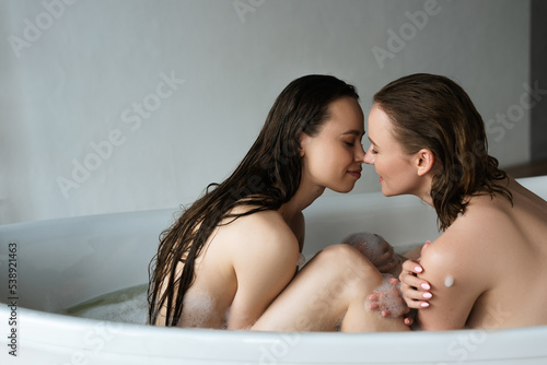 side view of naked lesbian women with closed eyes smiling face to face in bathtub.