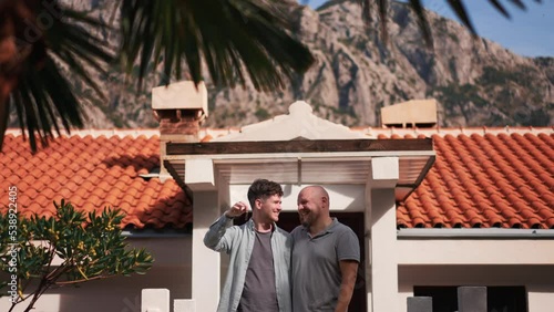 Beautiful young gay couple staying holding a key at background of new house. Two hapy homosexual men showing keys to new apartment lovely area on sunny day. Concept of Pride, lgbt family photo
