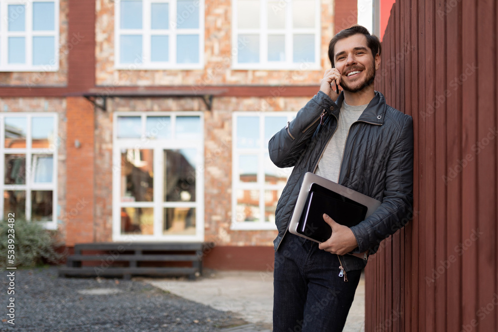 adult male businessman talking on a mobile phone leaning against a wooden fence next to a brick building