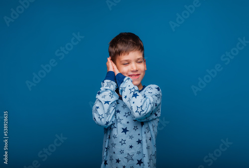Portrait of a cute little child standing with his hands closed, pretending to sleep isolated on a blue background. a boy in pajamas