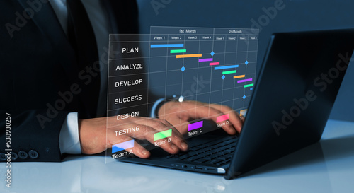  Project manager working on laptop and updating tasks and milestones progress planning with Gantt chart scheduling interface for company on virtual screen.Business Data Management System.