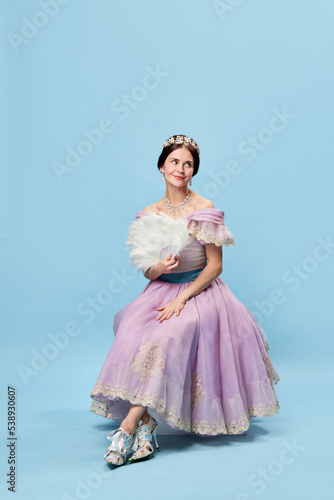 Shyness. Young elegant girl in lilac color medieval dress as young queen or princess on blue background. Eras comparison, beauty, art, emotions and vintage fashion style