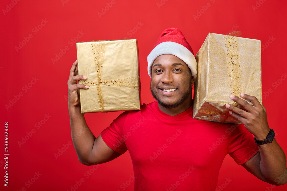 Portrait of a cute black guy with a toothy smile in a Santa hat, who holds Christmas gifts in golden packaging on his shoulders on a bright red background Portrait of a cute black guy with a toothy smile in a Santa hat, who holds Christmas gifts in golden packaging on his shoulders on a bright red background
