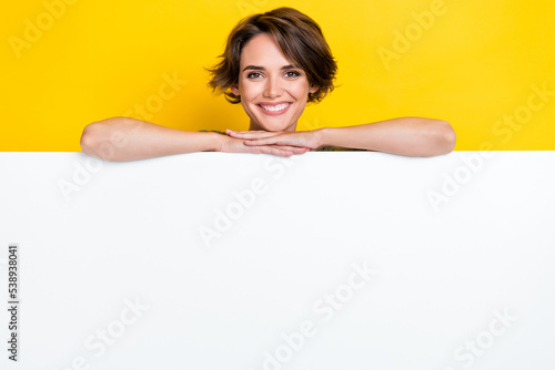 Photo of cheerful pretty cute lady hides behind empty blank space billboard recommend beauty procedure isolated on yellow color background