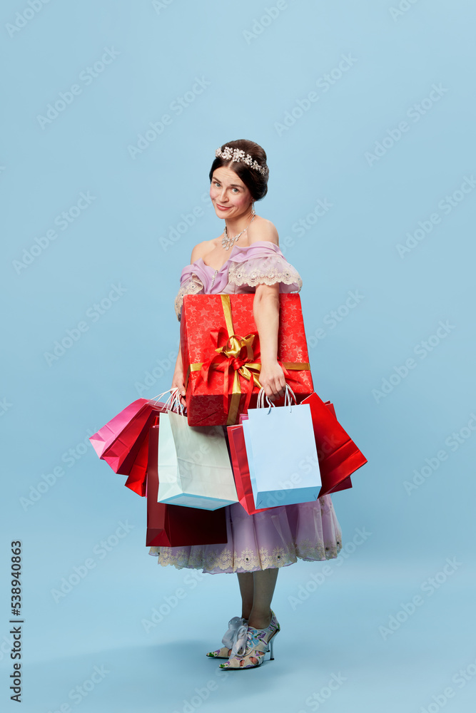 Great shopping day. Beautiful young woman in image of young queen or princess holding colorful shopping bags and festive boxes isolated on blue background.