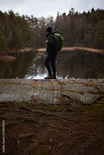 A caucasian man hiker standing on a rock over looking a lake in the forest watching reflections in the water with dramatic clouds in the sky.