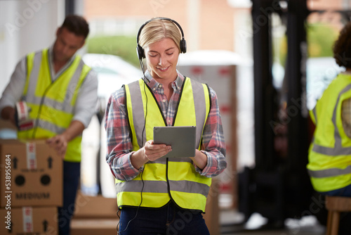 Female Worker Wearing Headset In Logistics Distribution Warehouse Using Digital Tablet