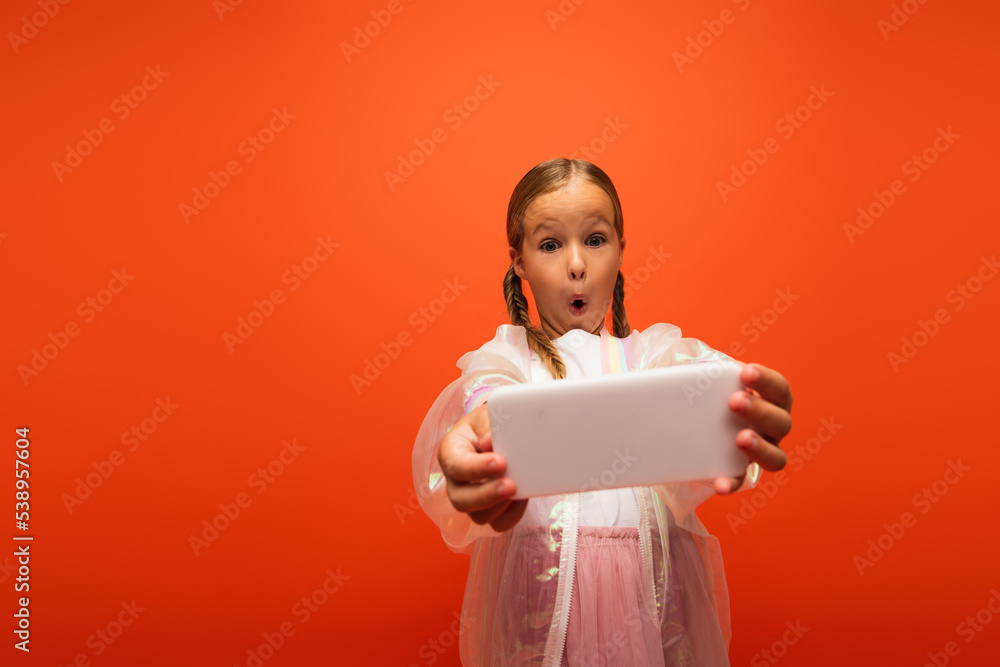 girl with open mouth and thrilled face expression taking selfie on cellphone isolated on orange girl with open mouth and thrilled face expression taking selfie on cellphone isolated on orange