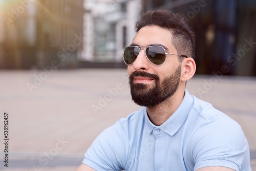 Portrait of happy positive bearded Arab Muslim ethnic guy, young man with black beard in black sunglasses and polo shirt is smiling outdoors at summer sunny day