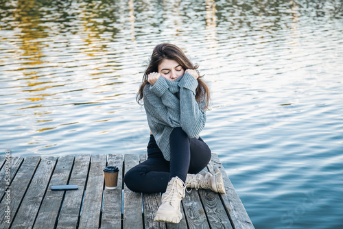 An attractive young woman in a sweater sits by the river. © puhimec