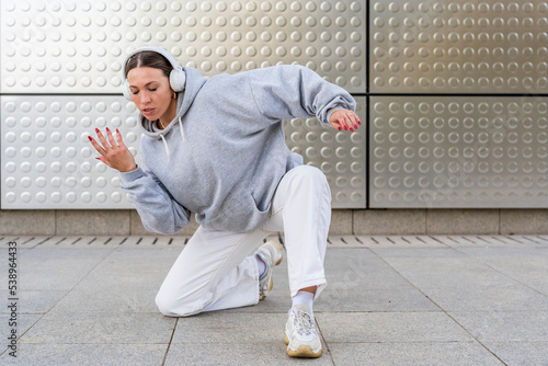 Young woman with headphones listening to urban music and dressed in white pants and gray hoodie dancing in front of metallic background urban dance