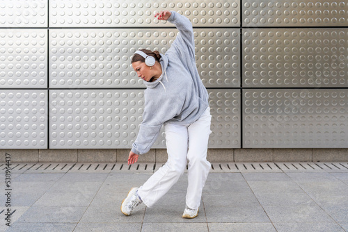 Young woman with headphones listening to urban music and dressed in white pants and gray hoodie dancing in front of metallic background urban dance