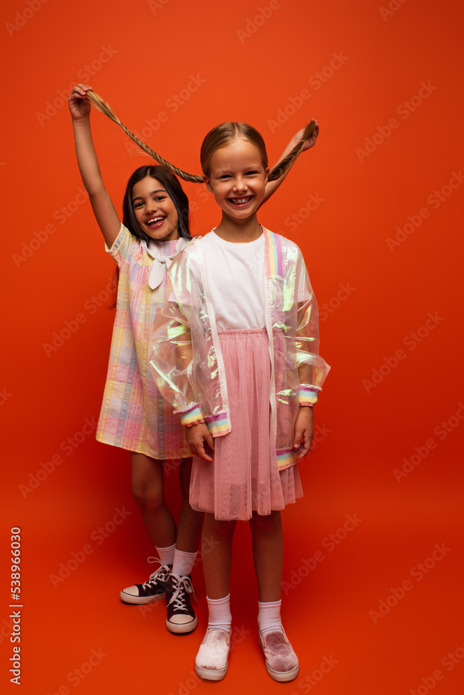 full length of excited girl holding pigtails of cheerful friend on orange background
