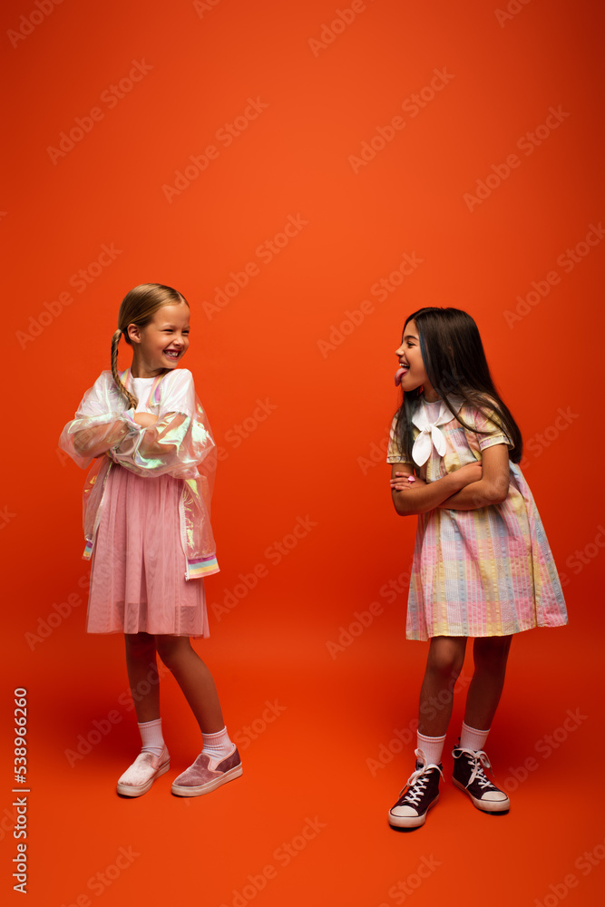 full length of brunette girl sticking out tongue to smiling friend standing with crossed arms on orange background