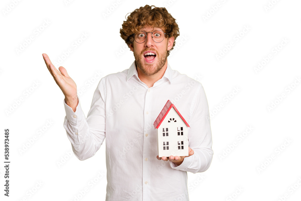 Business caucasian man holding toy house isolated on white background receiving a pleasant surprise, excited and raising hands. Business caucasian man holding toy house isolated on white background receiving a pleasant surprise, excited and raising hands.
