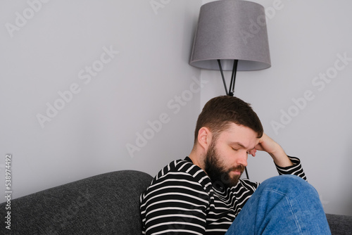 candid tired and sleepy beard man holding his head and sitting in the corner of the sofa on white background. Mental health, stressed day, headache and migraine