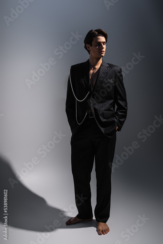 full length of man in trendy suit with pearls looking away while posing with hands in pockets on dark grey