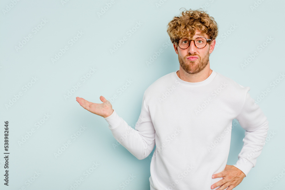Young caucasian man isolated on blue background showing a copy space on a palm and holding another hand on waist. Young caucasian man isolated on blue background showing a copy space on a palm and holding another hand on waist.