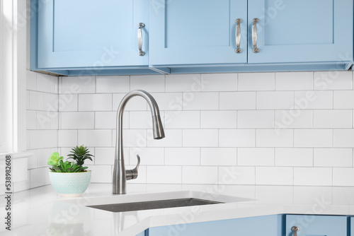 Detail of a kitchen with light blue cabinets, white granite countertop, subway tile backsplash, and a light hanging above a window.