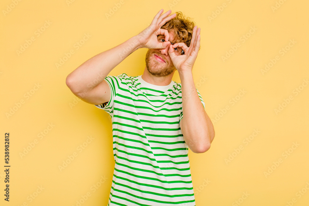 Young caucasian man isolated on yellow background showing okay sign over eyes Young caucasian man isolated on yellow background showing okay sign over eyes