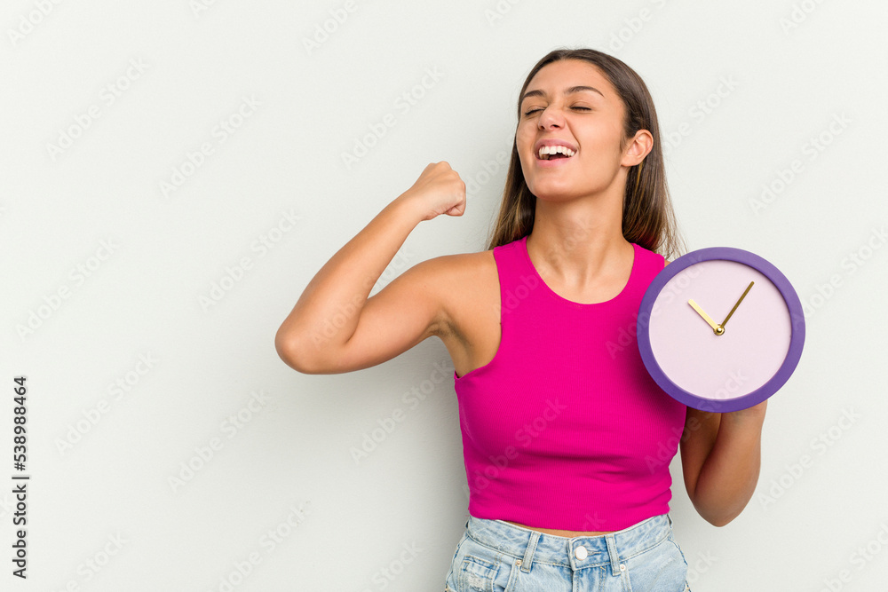 Young Indian woman holding a clock isolated on white background raising fist after a victory, winner concept. Young Indian woman holding a clock isolated on white background raising fist after a victory, winner concept.