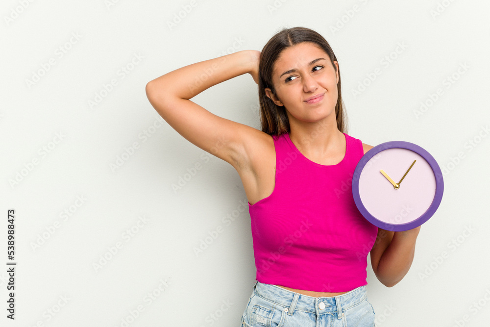 Young Indian woman holding a clock isolated on white background touching back of head, thinking and making a choice. Young Indian woman holding a clock isolated on white background touching back of head, thinking and making a choice.