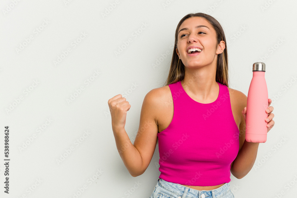 Young indian woman holding a pink thermo isolated on white background raising fist after a victory, winner concept. Young indian woman holding a pink thermo isolated on white background raising fist after a victory, winner concept.