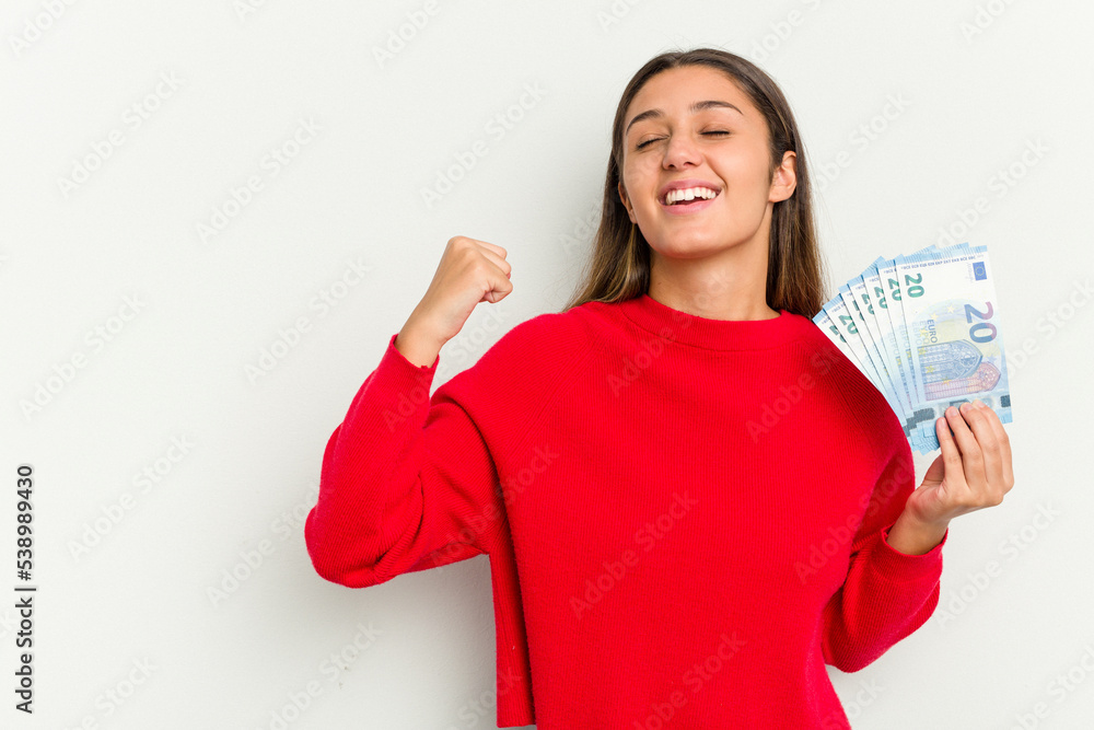 Young Indian woman holding a banknotes isolated on white background raising fist after a victory, winner concept. Young Indian woman holding a banknotes isolated on white background raising fist after a victory, winner concept.