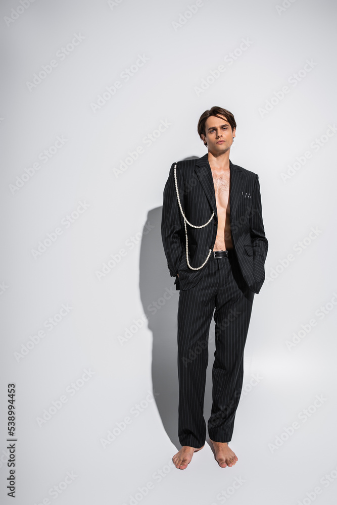 full length of barefoot and brunette man in trendy suit with pearls posing with hands in pockets on grey full length of barefoot and brunette man in trendy suit with pearls posing with hands in pockets on grey