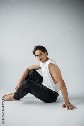 barefoot man in trousers and white t-shirt looking at camera while sitting on grey