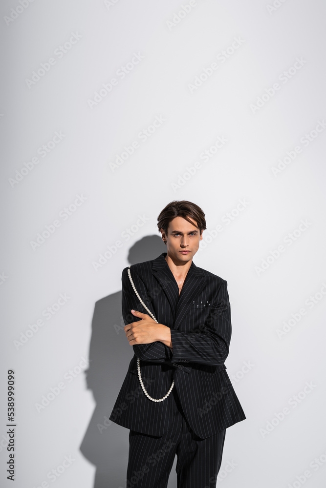 brunette man in trendy blazer with pearls posing with crossed arms while looking at camera on grey background brunette man in trendy blazer with pearls posing with crossed arms while looking at camera on grey background