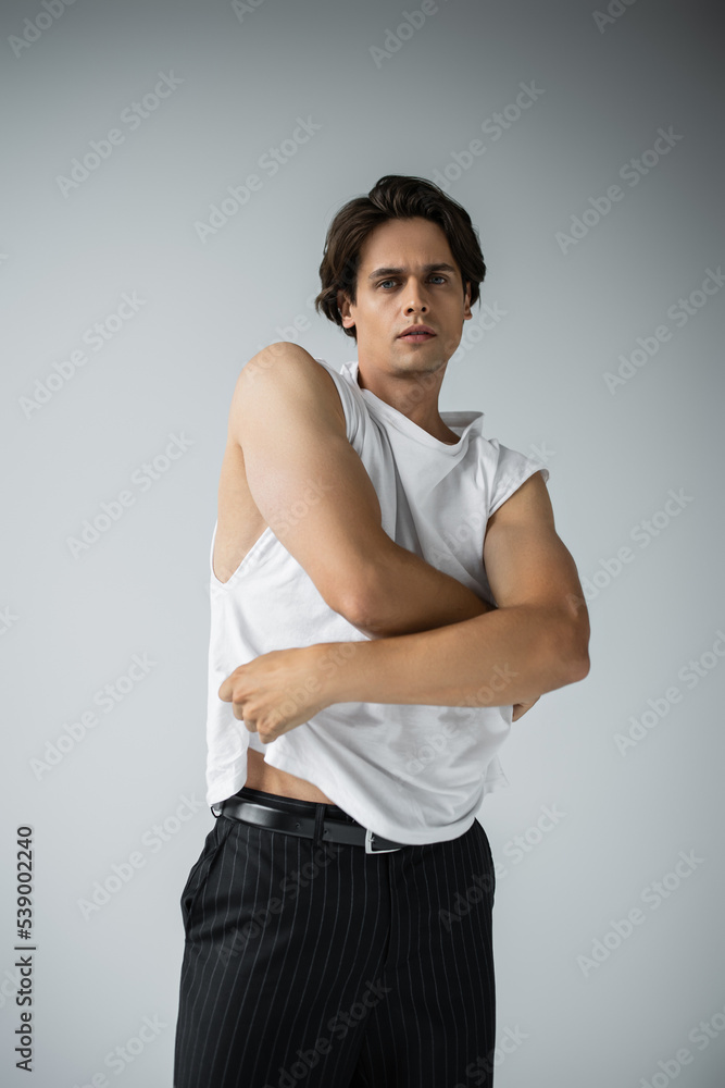 Obraz premium muscular man in trousers taking off white t-shirt and looking at camera on grey