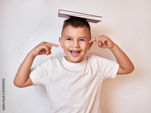 A little boy holds a book on his head with a finger raised.