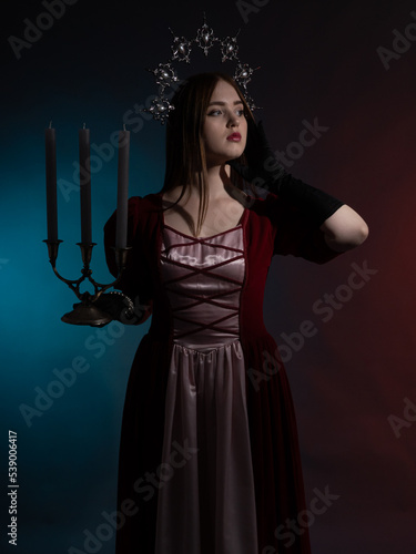 Mythical fantasy queen with a candlestick. Silvery gothic crown on the head. Burgundy vintage artistic dress, black gloves. Medieval style. Portrait in a dark key. Vertical photo.