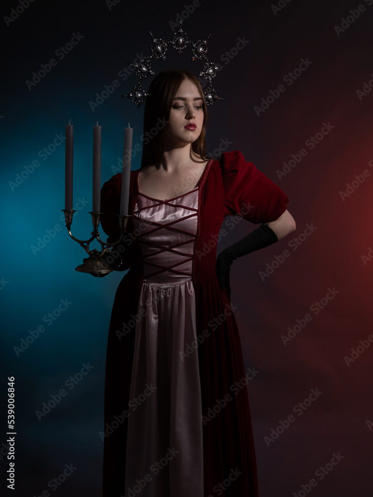 Mythical fantasy queen with a candlestick. Silvery gothic crown on the head. Burgundy vintage artistic dress, black gloves. Medieval style. Portrait in a dark key. Vertical photo.