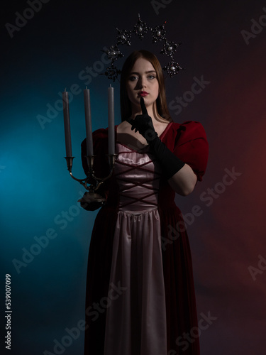 Mythical fantasy queen with a candlestick. Silvery gothic crown on the head. Burgundy vintage artistic dress, black gloves. Medieval style. Portrait in a dark key. Vertical photo.