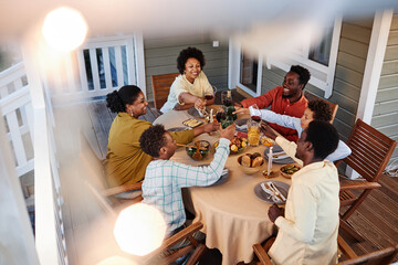High angle view at big African American family clinking glasses over dinner table outdoors in cozy setting with fairy lights