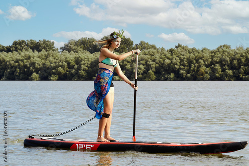 Young sexy woman in swimsuit and wreath on the head rowing on stand up paddle board. Water sports, active lifestyle.