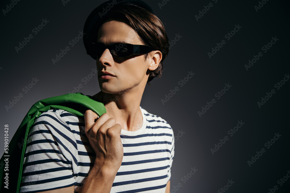 Obraz premium Stylish man in sunglasses holding green leather jacket isolated on grey with lighting